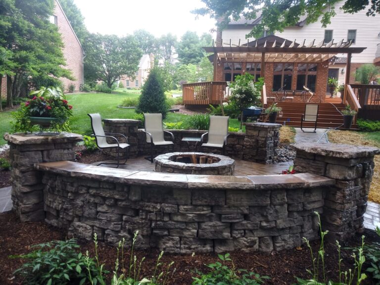 Stacked stone seating wall and fire pit patio with a pergola-covered deck and brick home beyond.