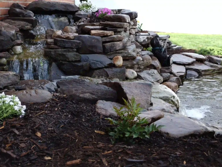 Stacked stone waterfall spilling into a backyard pond, with boulders, dark mulch and pink and white blooms