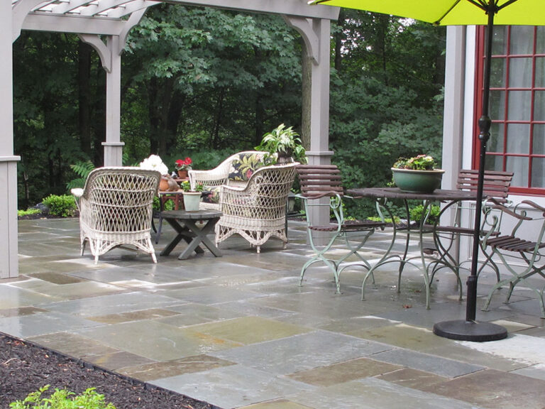 Bluestone patio with wicker chairs, bistro dining set, and a green umbrella against a woodland backdrop.