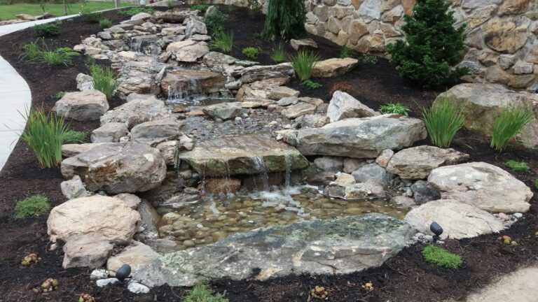 Pondless boulder stream with multiple small waterfalls, grasses and landscape lighting beside a stone wall in York PA