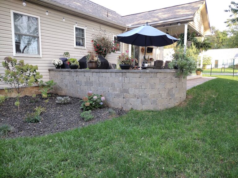 Curved block retaining wall supporting a raised patio with umbrella seating, with new flowering shrub and perennial beds below