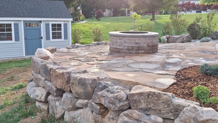 Flagstone patio and round block fire pit on a boulder-edged terrace beside a gray backyard shed.