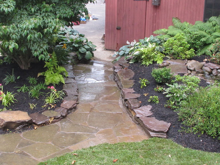 Wet flagstone walkway winding between mulched beds of shade plantings beside a red barn.