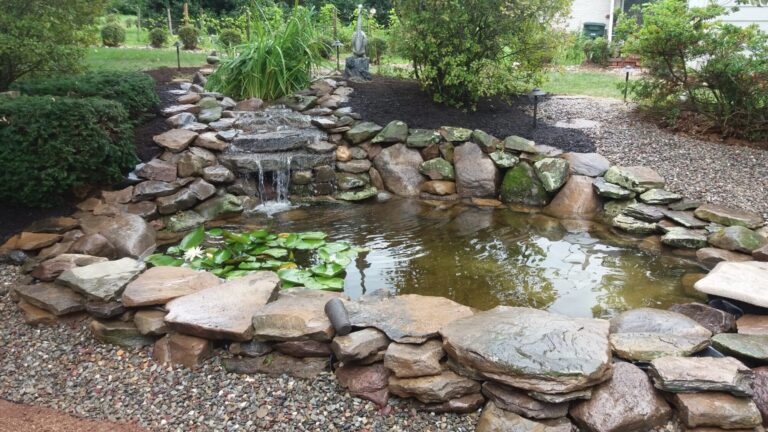 Rock-edged garden pond with waterfall, aquatic plants and a heron statue surrounded by fresh mulch beds