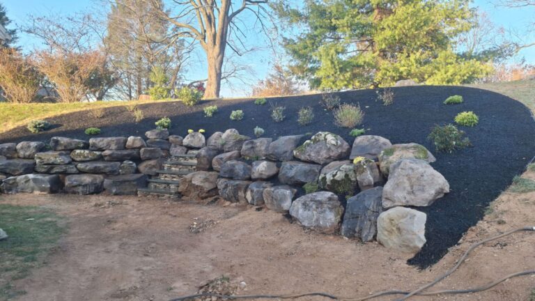 Large boulder retaining wall holding back a mulched hillside, with built-in stone steps leading up through the wall