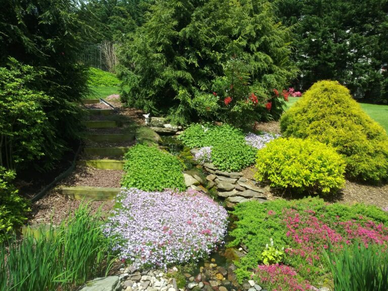 Sloped rock garden with flowering groundcover, golden shrubs and timber steps beside a small waterfall in York PA