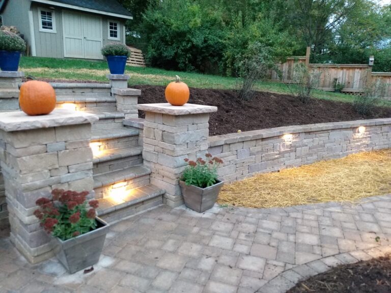 Stone steps and pillars with built-in lighting and pumpkins above a lighted retaining wall on a paver patio.