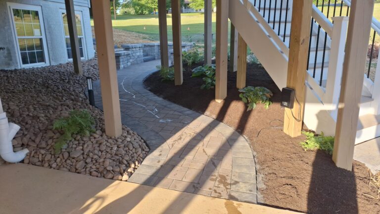 Curved paver walkway under an elevated deck with river rock, young shade plantings, and landscape lighting.