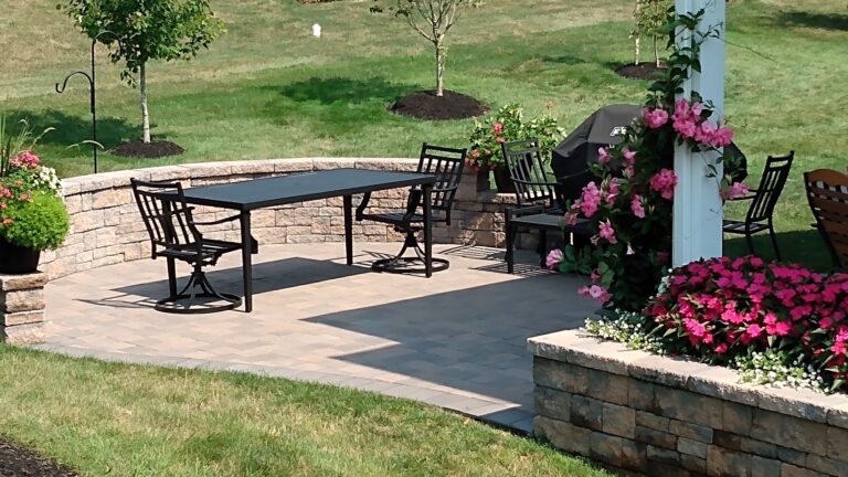 Paver patio with curved block sitting wall, dining table, and a pink flowering vine climbing a white pergola post.