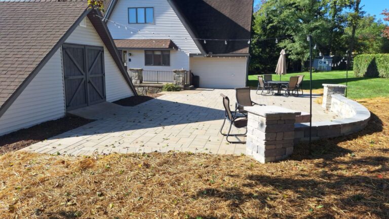 Light paver patio with stacked stone pillars, curved seat wall and overhead string lights connecting an A-frame home and barn