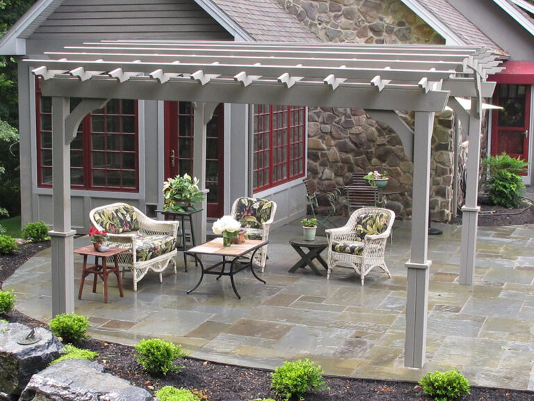 Painted pergola shading a bluestone patio with wicker furniture beside a stone cottage in York PA.