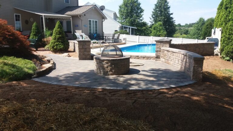 Paver patio with block fire pit, curved seating wall and pillars beside a fenced inground pool in York PA