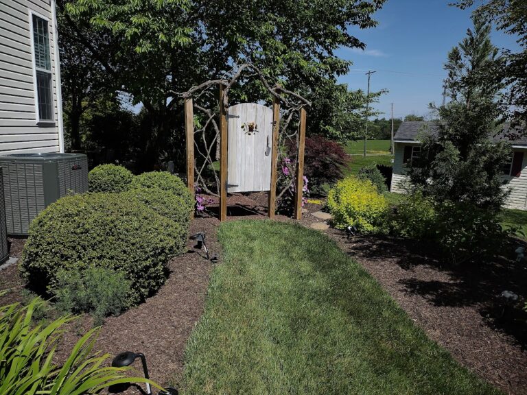 Whitewashed garden gate under a rustic branch arbor at the end of a grass path lined with evergreen shrubs in a York PA backyard