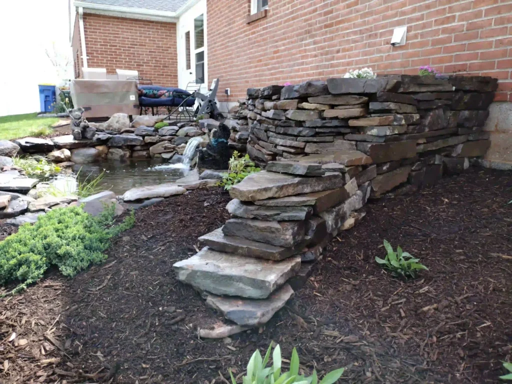 Dry-stacked stone retaining wall and flagstone steps beside a small pond and waterfall at a brick home in York PA