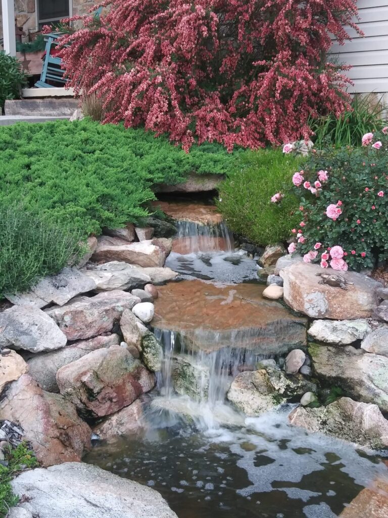 Close-up of water cascading over layered stone beside pink flowers and a small bronze frog statue.