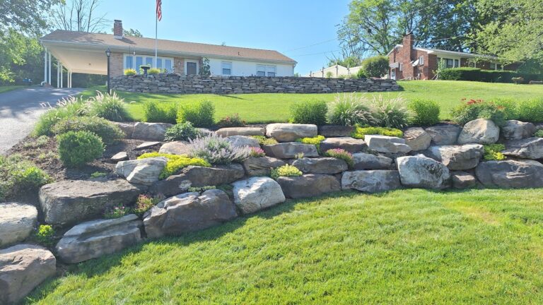 Terraced boulder retaining wall with perennials and ornamental grasses below a stone-walled ranch home in York PA.