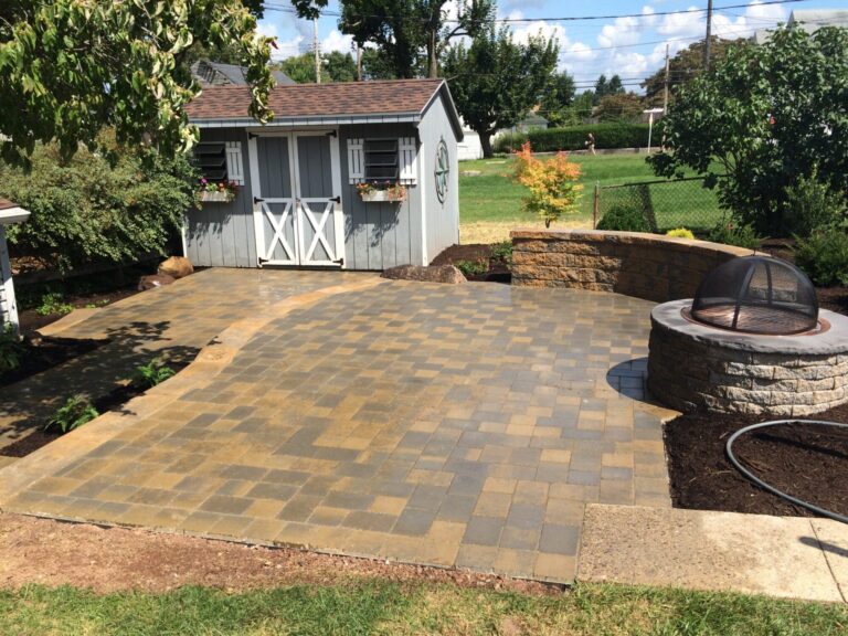 Tan and gray paver patio beside a gray shed, with a round stone fire pit with grill screen and curved retaining wall